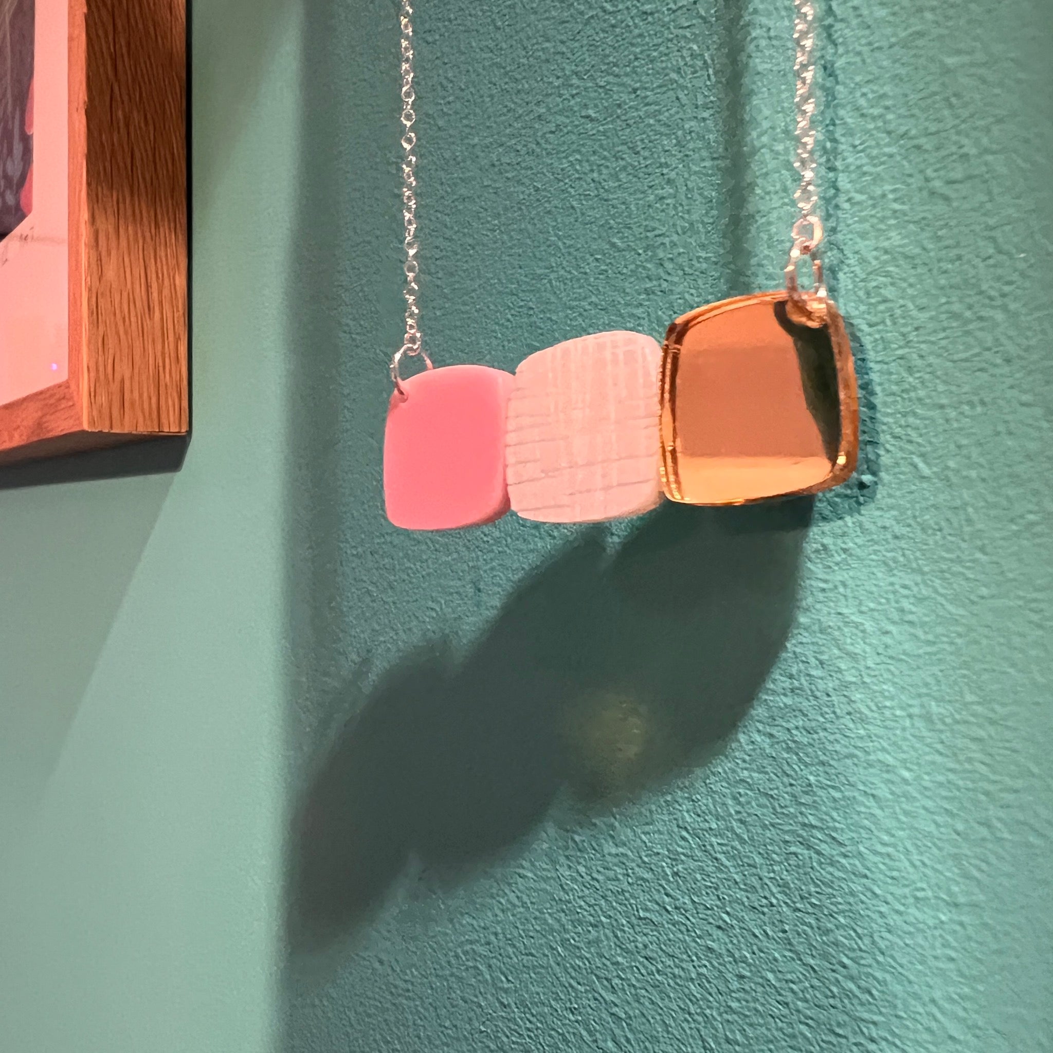 A gold pink and frosted white acrylic necklace featuring a trio of abstract pebble shapes. Seen hanging in front of a teal-coloured painted wall.