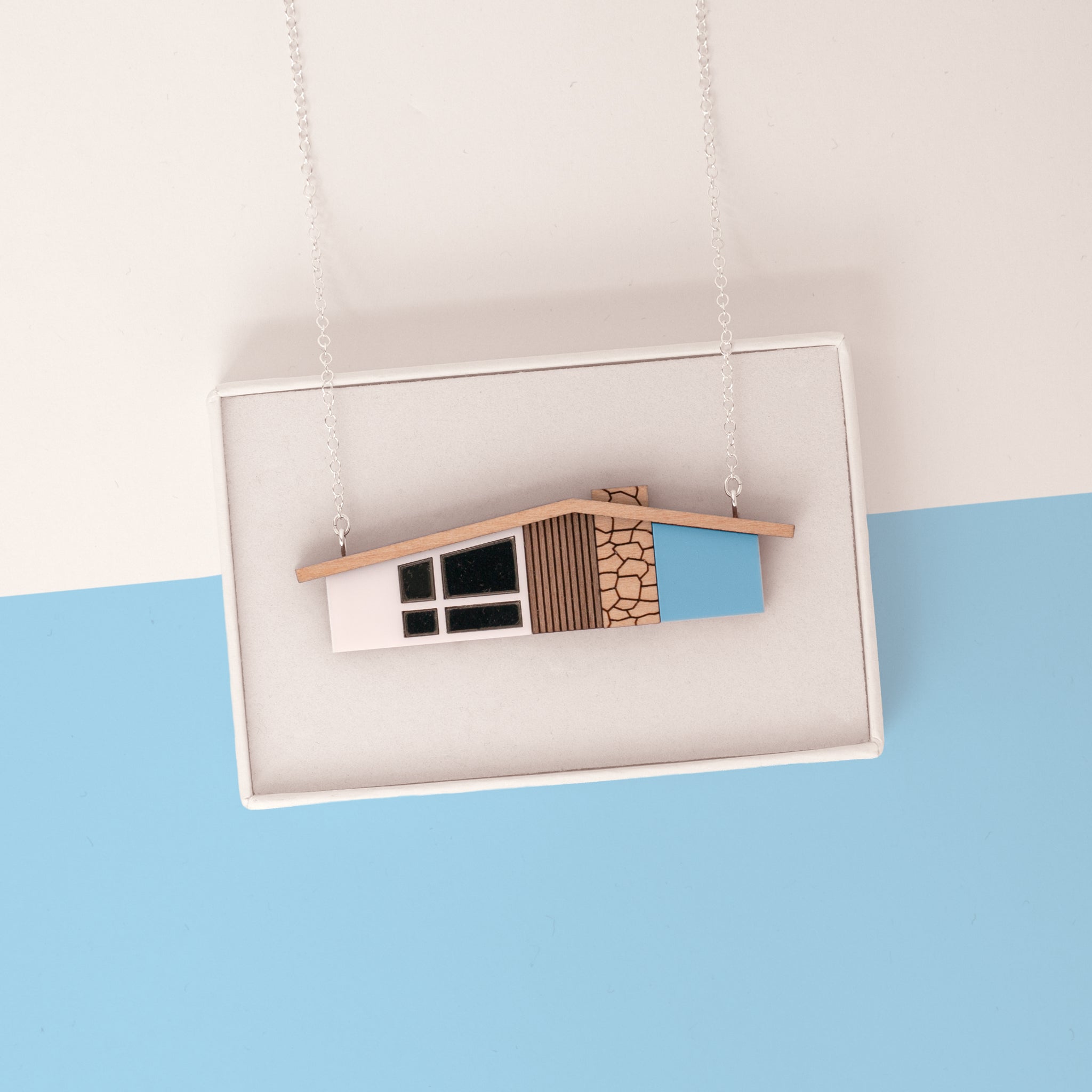 Blue midcentury house necklace on silver chain draped over a white gift box on a light blue and white background