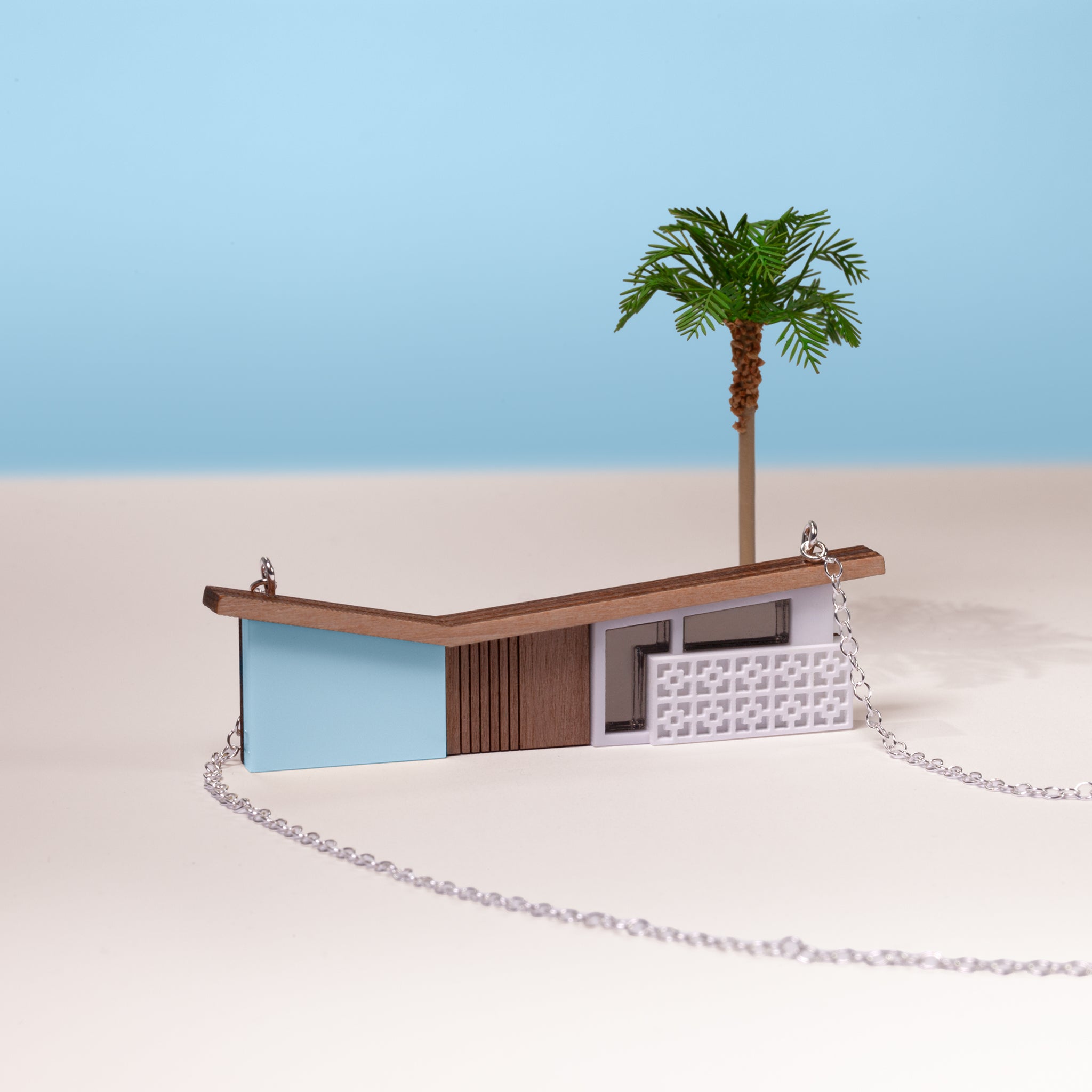 Blue house-shaped necklace on silver chain with a miniature model of a palm tree on a white and blue background