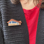 A close-up of a person wearing a wooden coffee-shop-themed brooch on a dark gray knit cardigan over a red t-shirt.