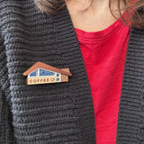 A close-up of a person wearing a wooden coffee-shop-themed brooch on a dark gray knit cardigan over a red t-shirt.