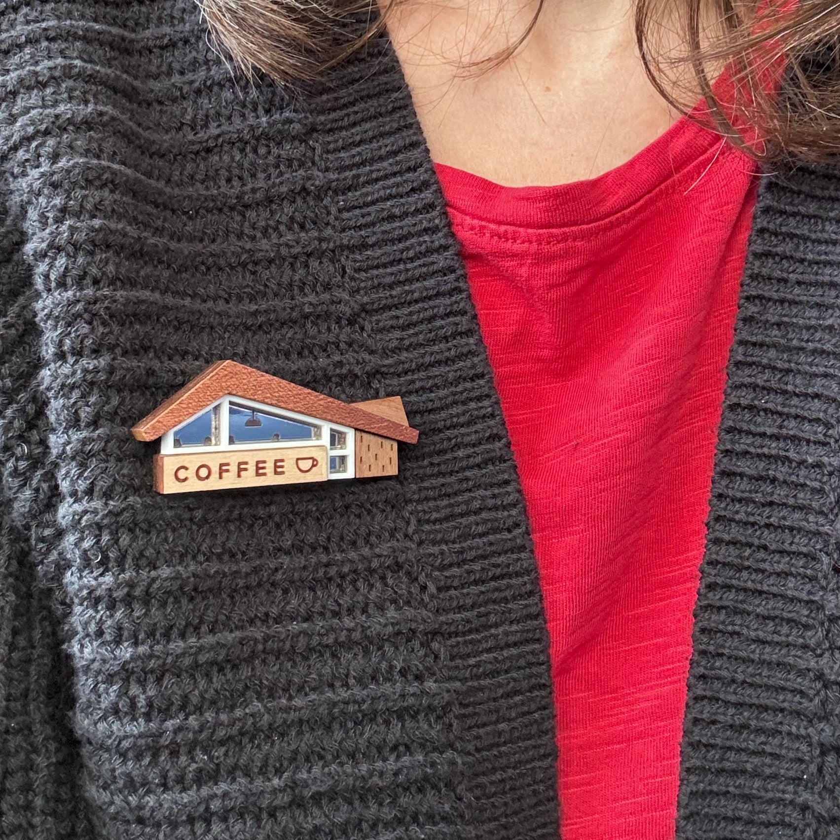 A close-up of a person wearing a wooden coffee-shop-themed brooch on a dark gray knit cardigan over a red t-shirt.