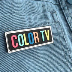 A typographic brooch with rainbow lettering on a blue denim jacket. The lettering reads 'COLOR TV'.