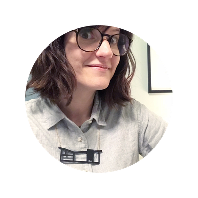 A circular photo of Jo from Tiny Scenic - a woman with dark hair and round glasses wearing a light gray button-up shirt, shown in a circular portrait crop against a white background.