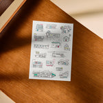 A white rectangular art print featuring black ink drawings of small storefronts and playful signs, with a few coloured accents, placed on a warm brown wooden surface.