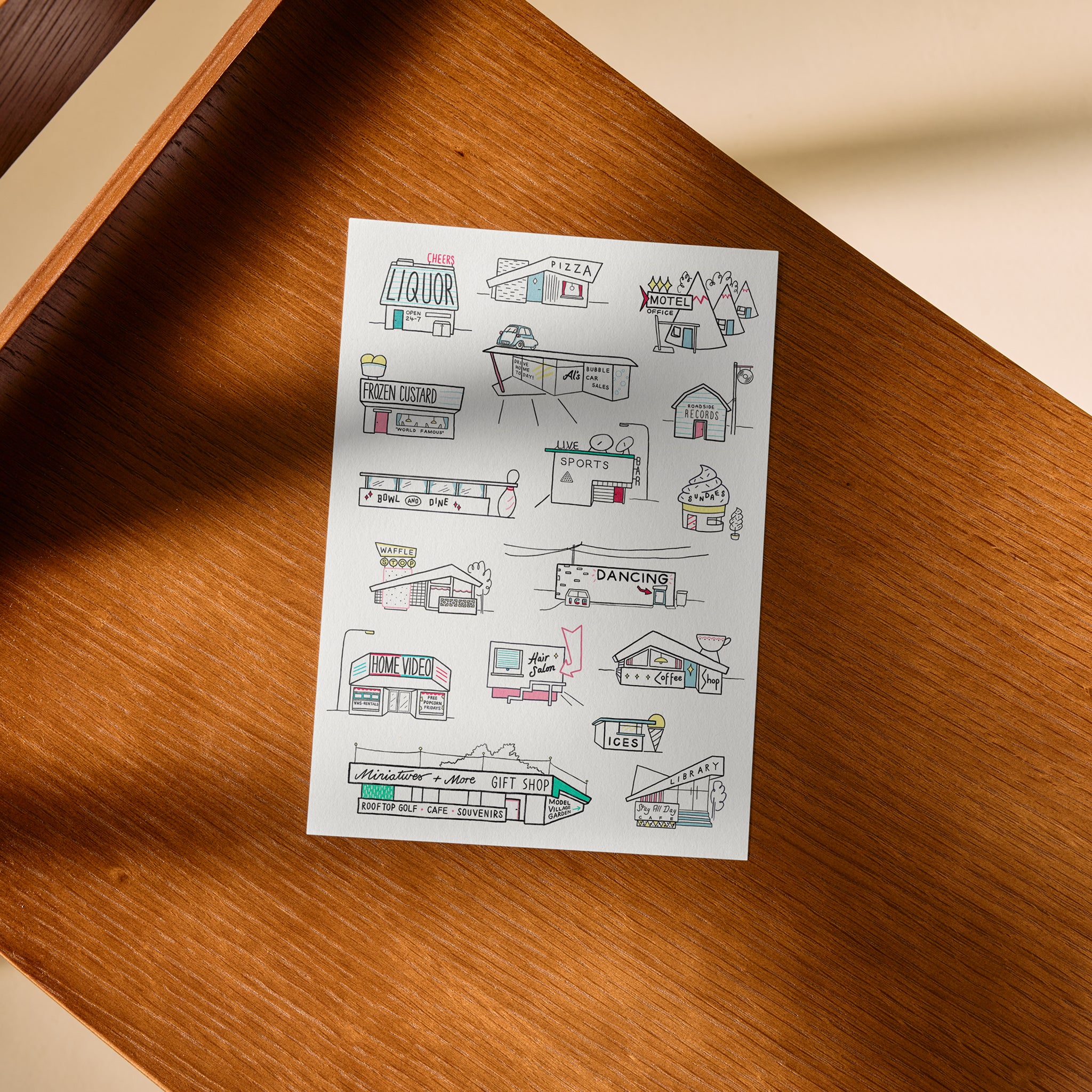 A white rectangular art print featuring black ink drawings of small storefronts and playful signs, with a few coloured accents, placed on a warm brown wooden surface.