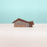 A front-on view of a miniature coffee shop necklace, handmade in wood and acrylic with sterling silver chain. Set against a white and blue background. The design of the coffee shop is inspired by googie architecture.