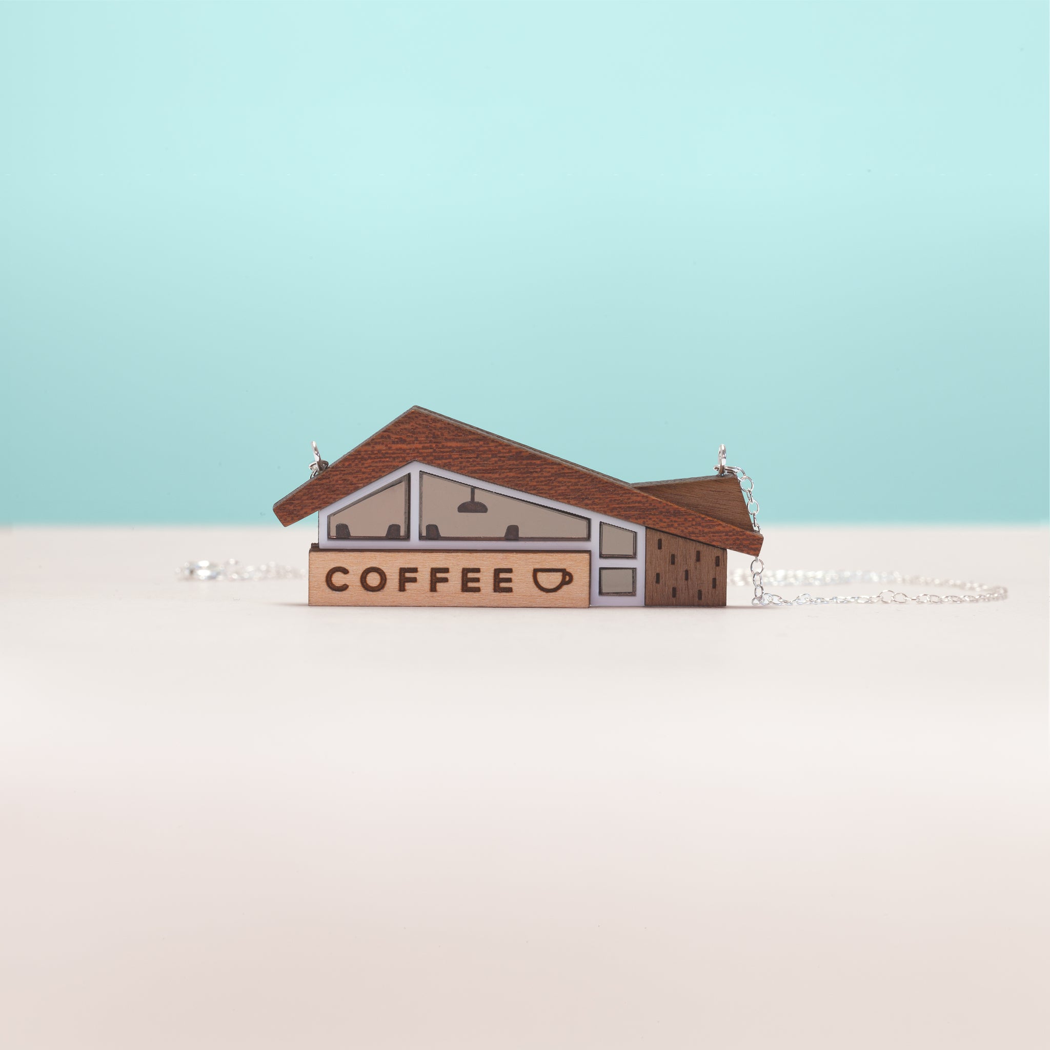 A front-on view of a miniature coffee shop necklace, handmade in wood and acrylic with sterling silver chain. Set against a white and blue background. The design of the coffee shop is inspired by googie architecture.