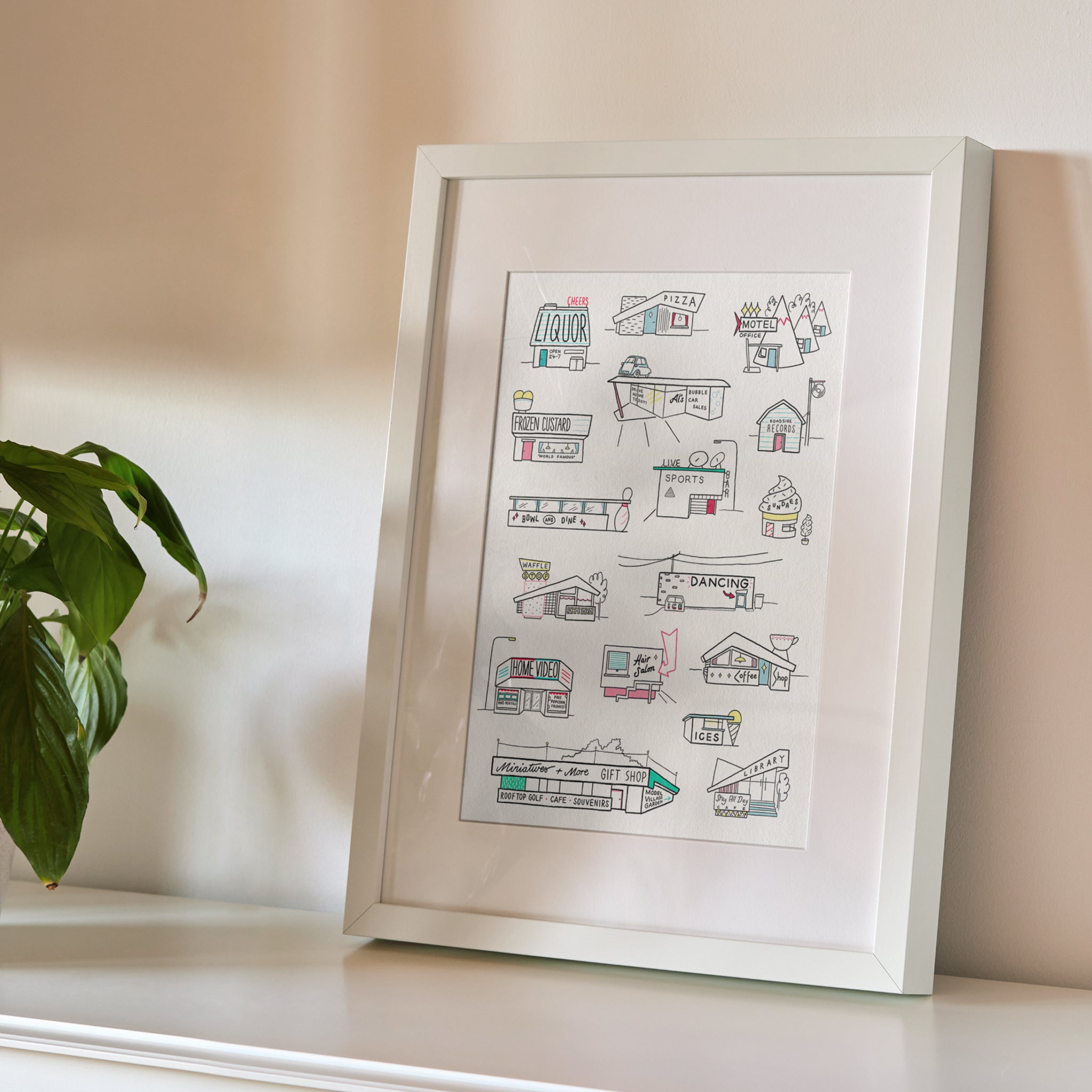 A framed art print featuring hand-drawn line art illustrations of retro roadside buildings and storefronts. Styled on a white shelf next to a green plant against a neutral beige wall.