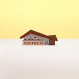 A miniature midcentury-era coffee shop brooch made from wood and acrylic, set against a plain white and yellow background.