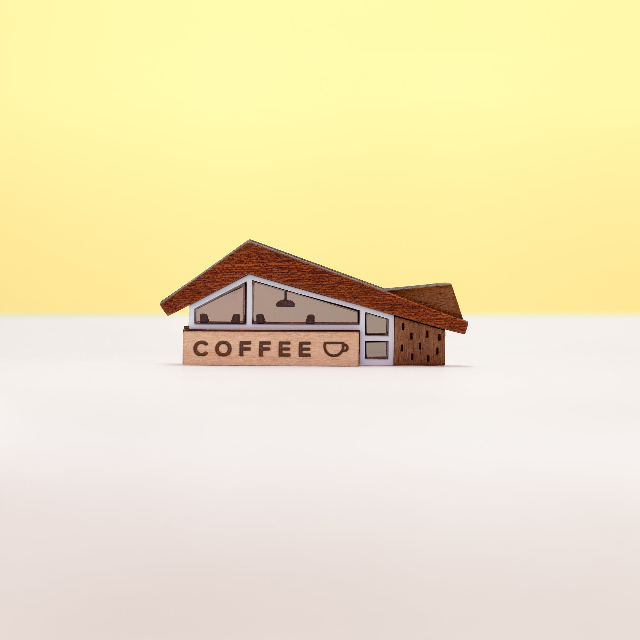 A miniature midcentury-era coffee shop brooch made from wood and acrylic, set against a plain white and yellow background.