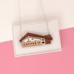 A wooden coffee shop house pendant necklace displayed inside a white presentation box. The pendant features a brown wood-grain roof, a light wood sign reading 'COFFEE' with a small heart, and white and dark brown window details, suspended from a thin silver chain against a pink and white background.