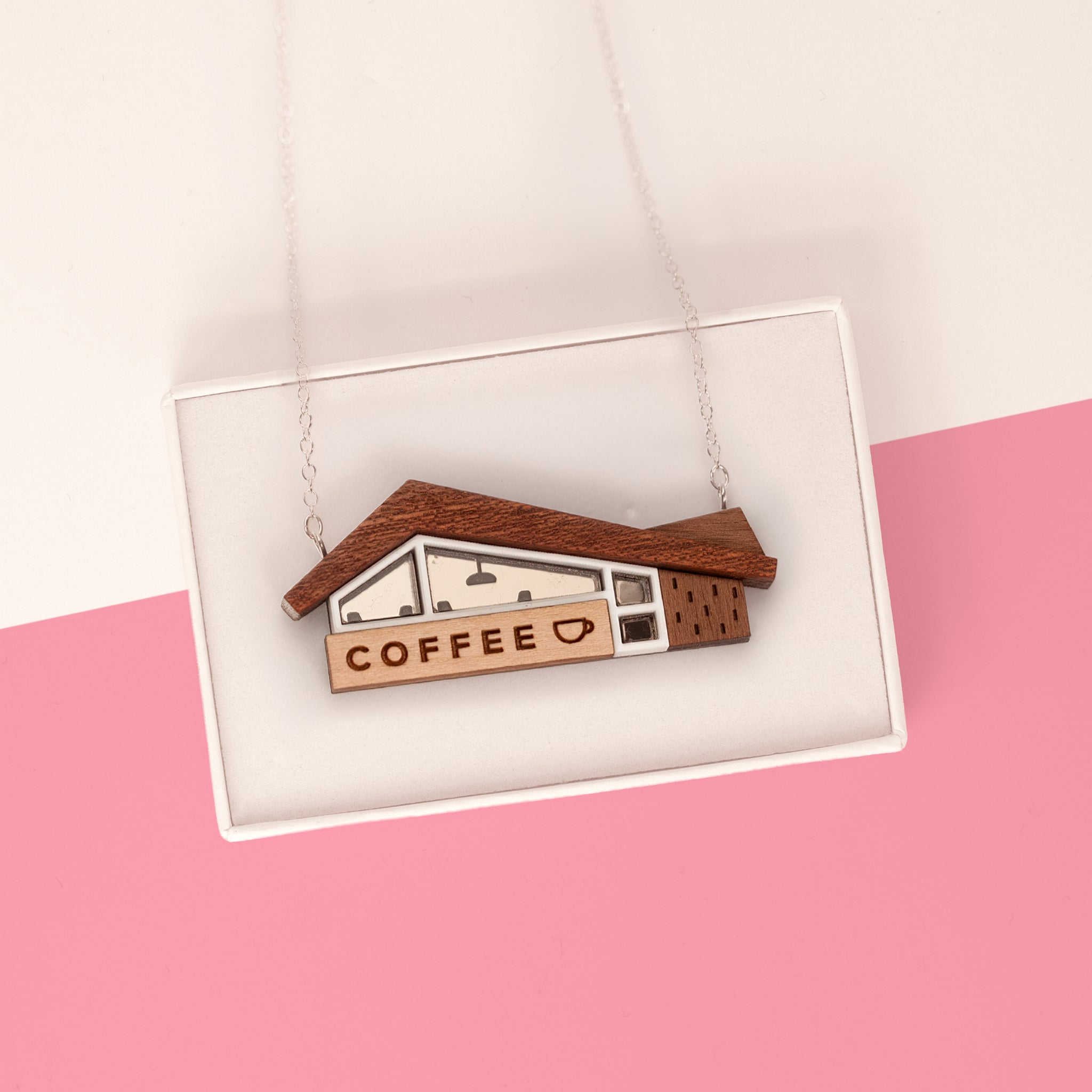 A wooden coffee shop house pendant necklace displayed inside a white presentation box. The pendant features a brown wood-grain roof, a light wood sign reading 'COFFEE' with a small heart, and white and dark brown window details, suspended from a thin silver chain against a pink and white background.