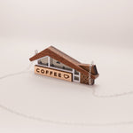 A miniature coffee shop pendant necklace made from wood and acrylic, seen from a side angle. The coffee shop has a retro Googie archictecture aesthetic.