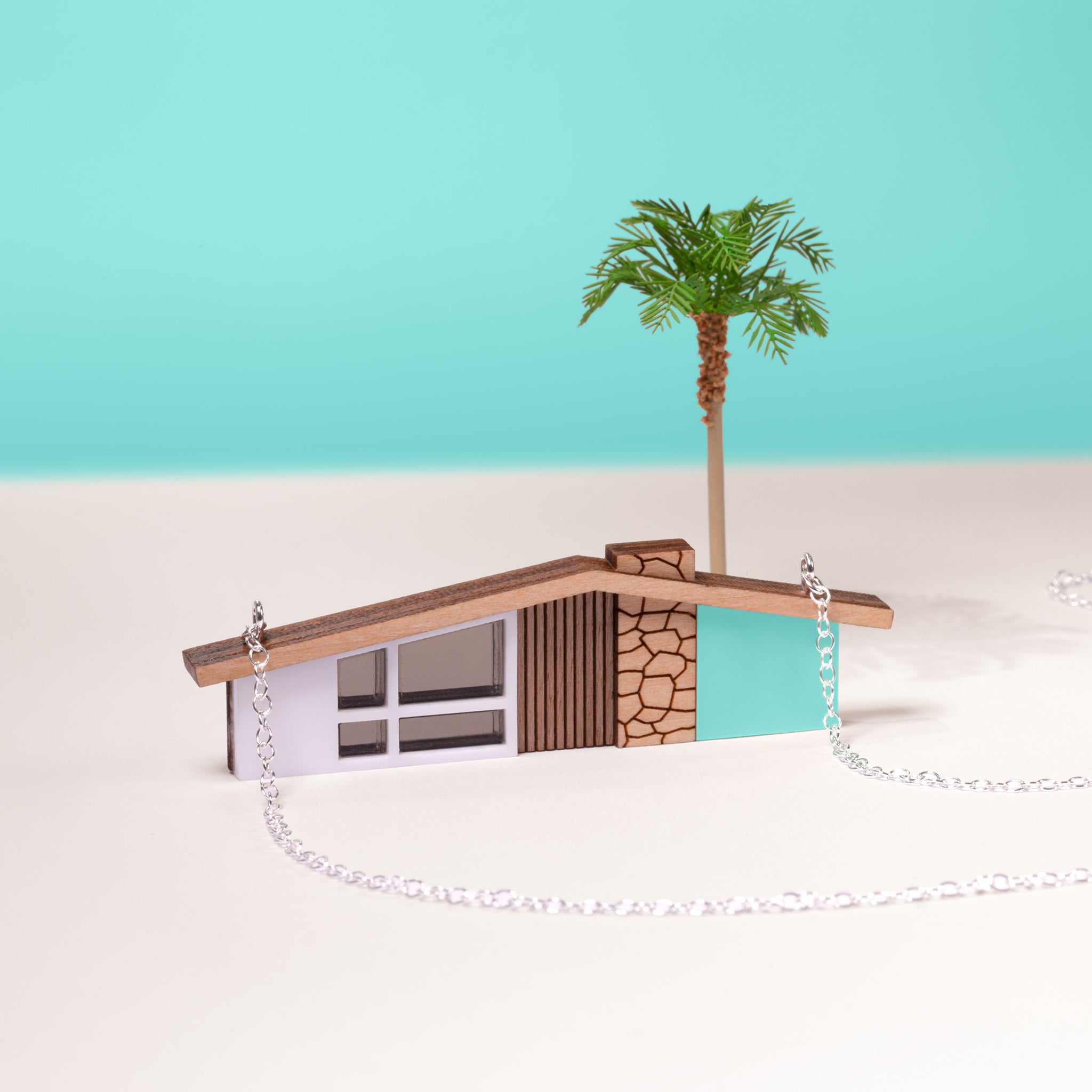 Miniature house necklace on a mint and white background with a miniature model of a palm tree