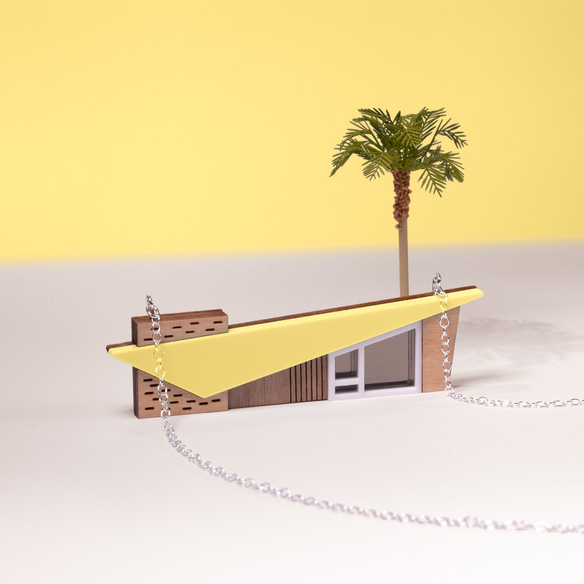 Colourful yellow retro midcentury house necklace with a miniature palm tree behind set on a white and yellow minimalist background