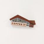 A wood and acrylic miniature coffee shop novelty brooch with teeny tiny lighting and diner booth seating details in the windows and a coffee cup illustration on the sign. The brooch is set against a plain white background. 