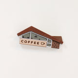 A wood and acrylic miniature coffee shop novelty brooch with teeny tiny lighting and diner booth seating details in the windows and a coffee cup illustration on the sign. The brooch is set against a plain white background. 