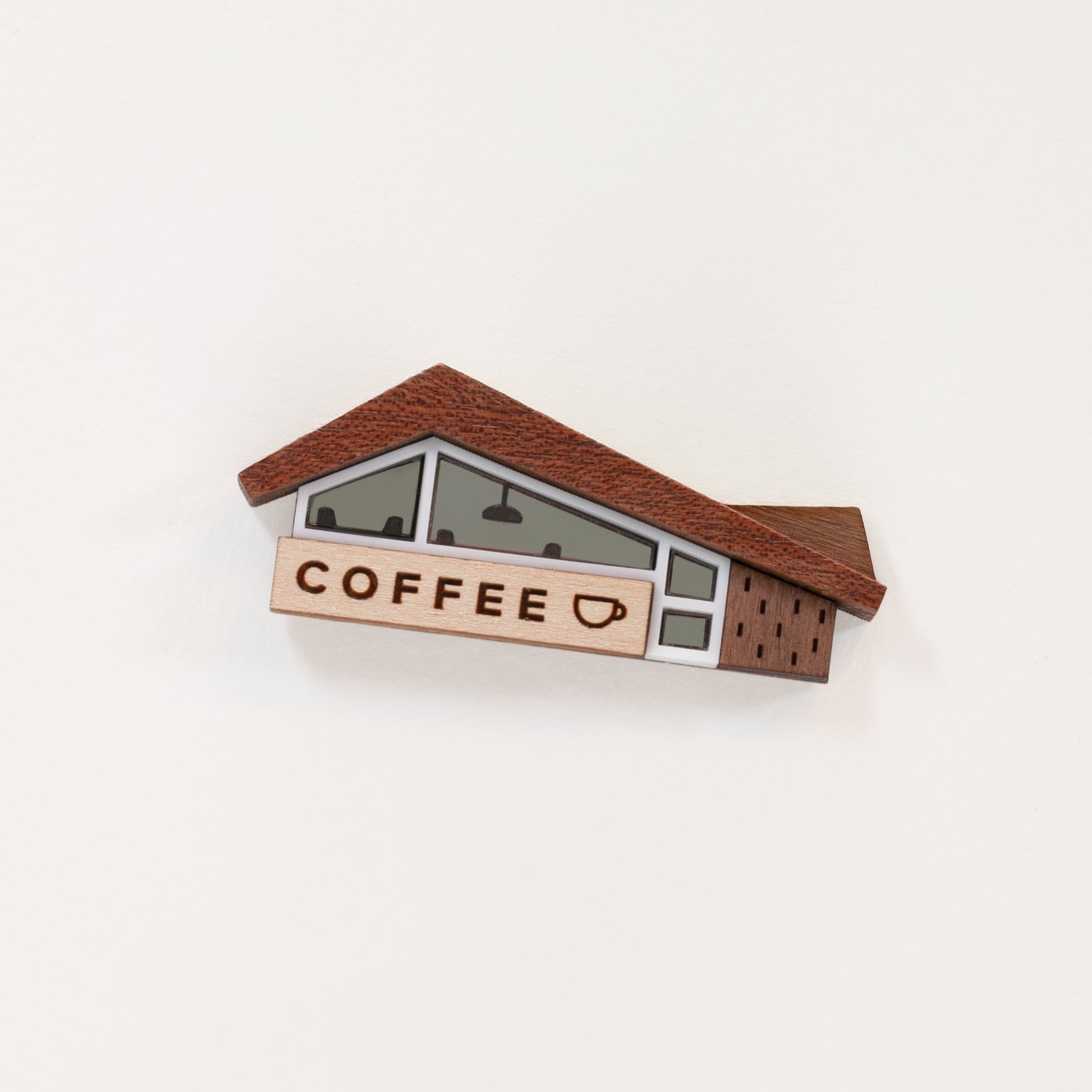 A wood and acrylic miniature coffee shop novelty brooch with teeny tiny lighting and diner booth seating details in the windows and a coffee cup illustration on the sign. The brooch is set against a plain white background. 