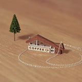 Close-up detail of retro wooden coffee shop pendant showing the coffee sign and window details. The necklace stands on a wooden background in a scene with a miniature tree.