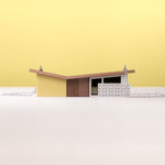 A miniature model of a yellow modernist house on silver chain against a yellow background