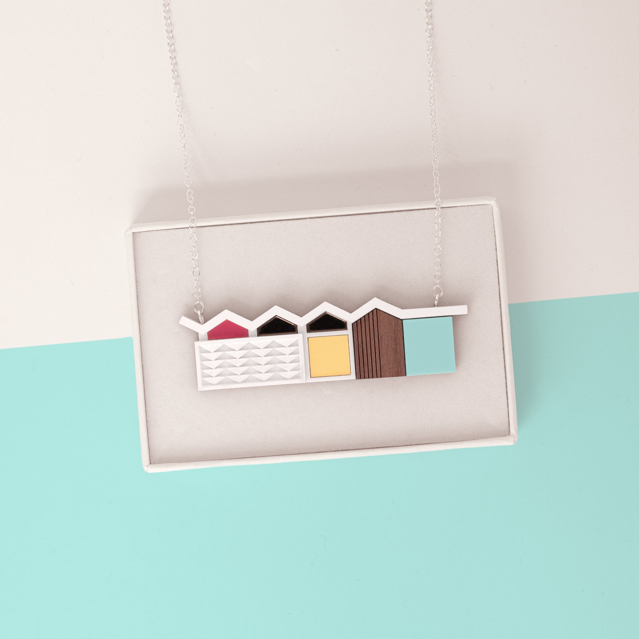 Colourful house pendant with zigzag roof draped over a white gift box on a light blue and white minimalist background